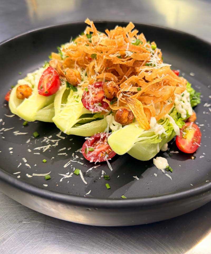 Sesión 5 (Clásicos reinterpretados y emplatado libre):
Ensalada César sin huevo. Cogollos de lechuga romana, aderezo César con aquafaba (agua de cocción de garbanzos), tiras crujientes de pasta oriental, queso parmesano, jitomate cherry, chips de ajo.